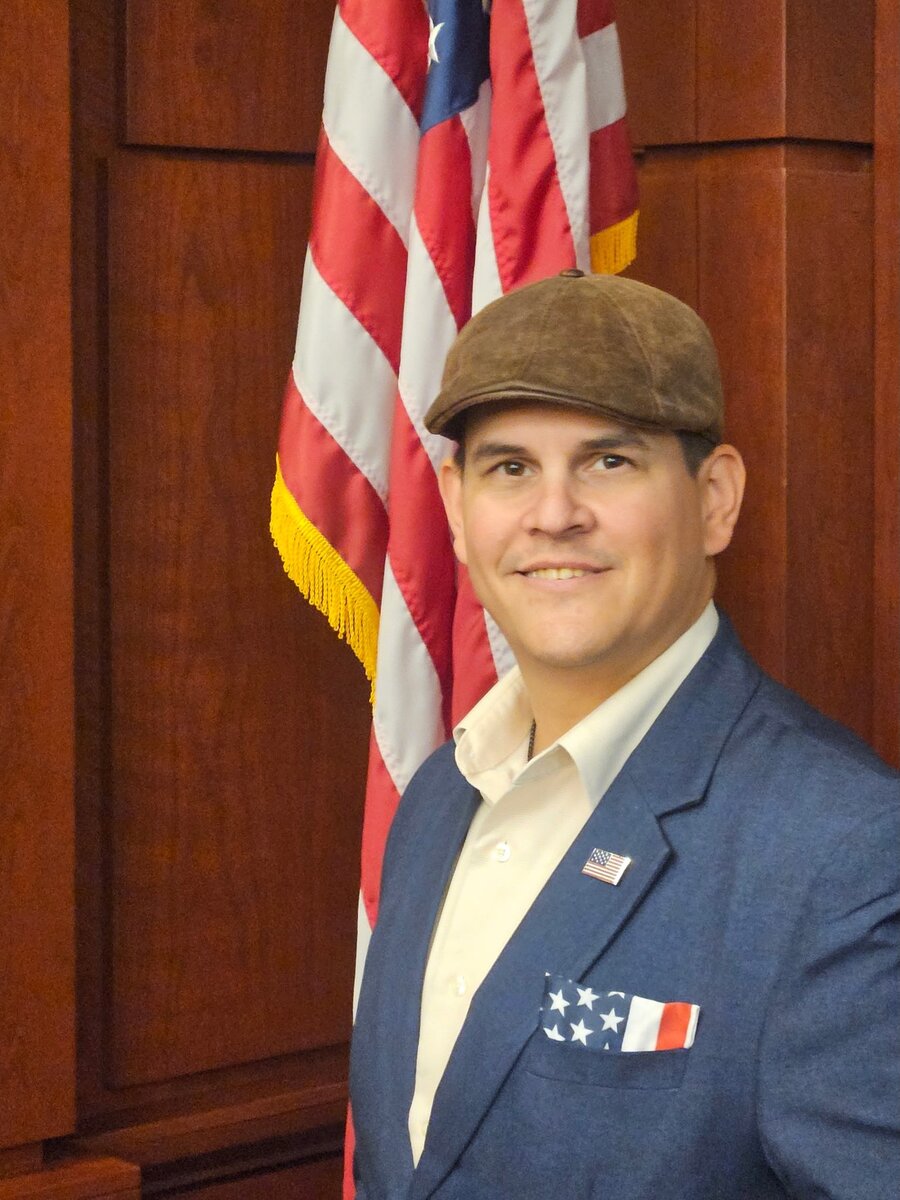 Tim Cortrecht in a flat cap and navy blazer with an American flag lapel pin, standing before a flag.